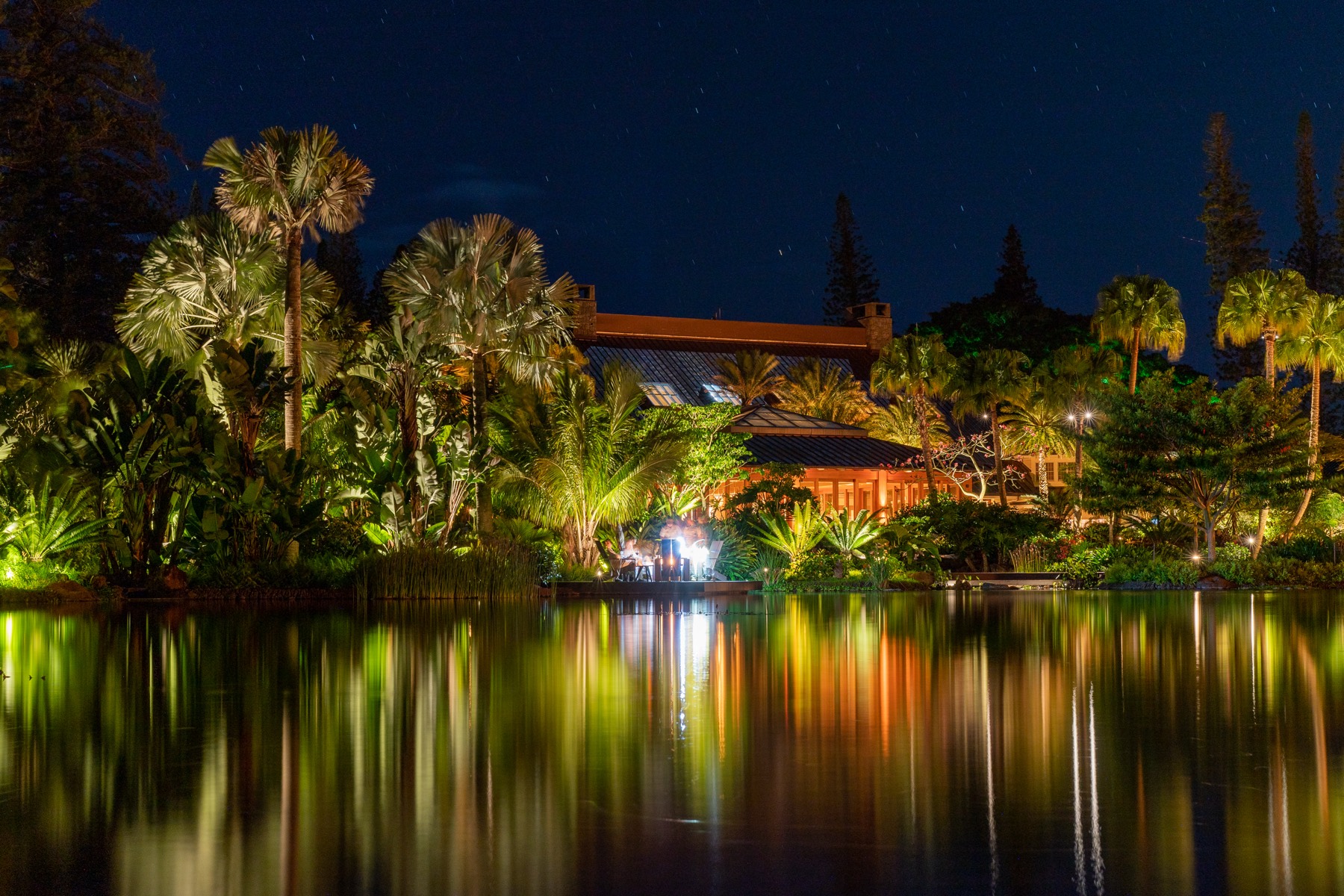 A resort lit at night, reflected in still water among palms.