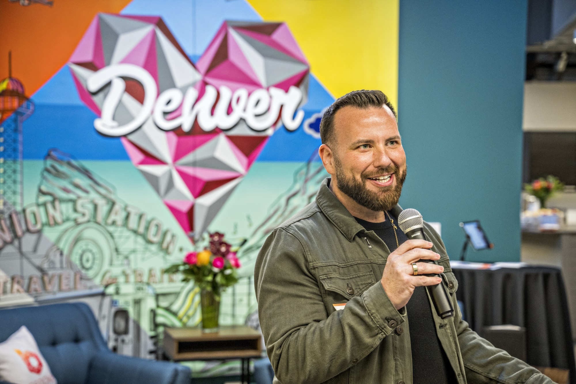 Justin speaking into a microphone in front of a colourful Denver mural.